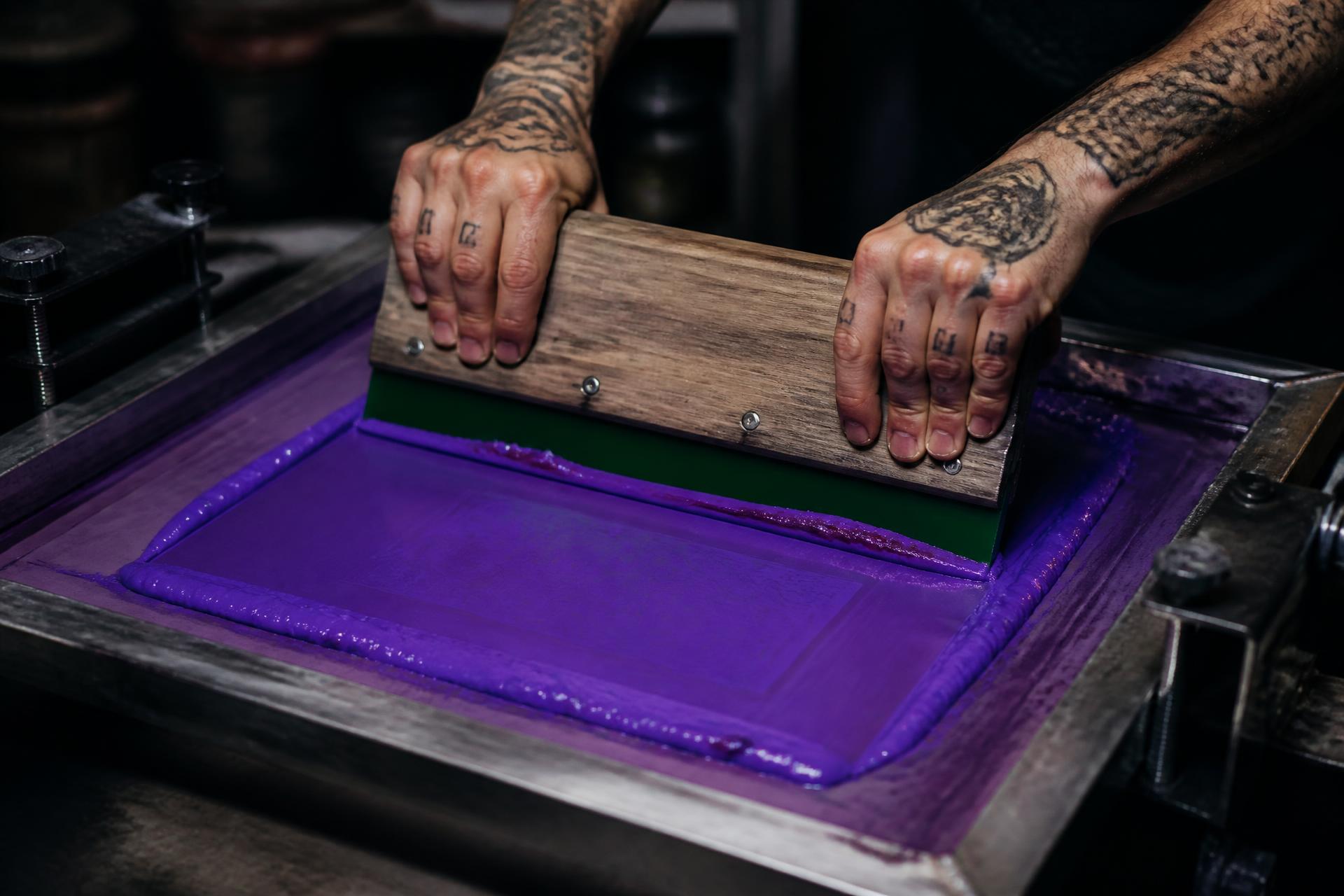 Manos tatuadas pasando la espátula con tinta morada sobre una pantalla de serigrafía